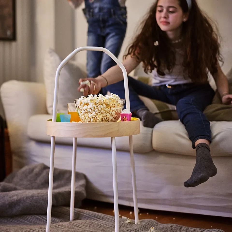 Two girls on sofa, one with remote, popcorn around, colourful containers on table.