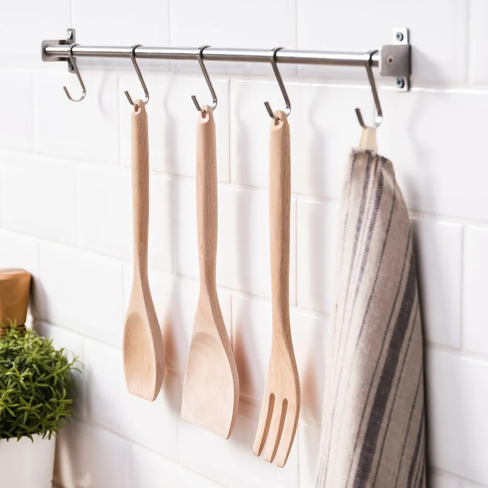 Six wooden cooking utensils hang from a stainless steel rail on a tiled wall, including a spoon, spatula, and fork, along with a striped tea towel.