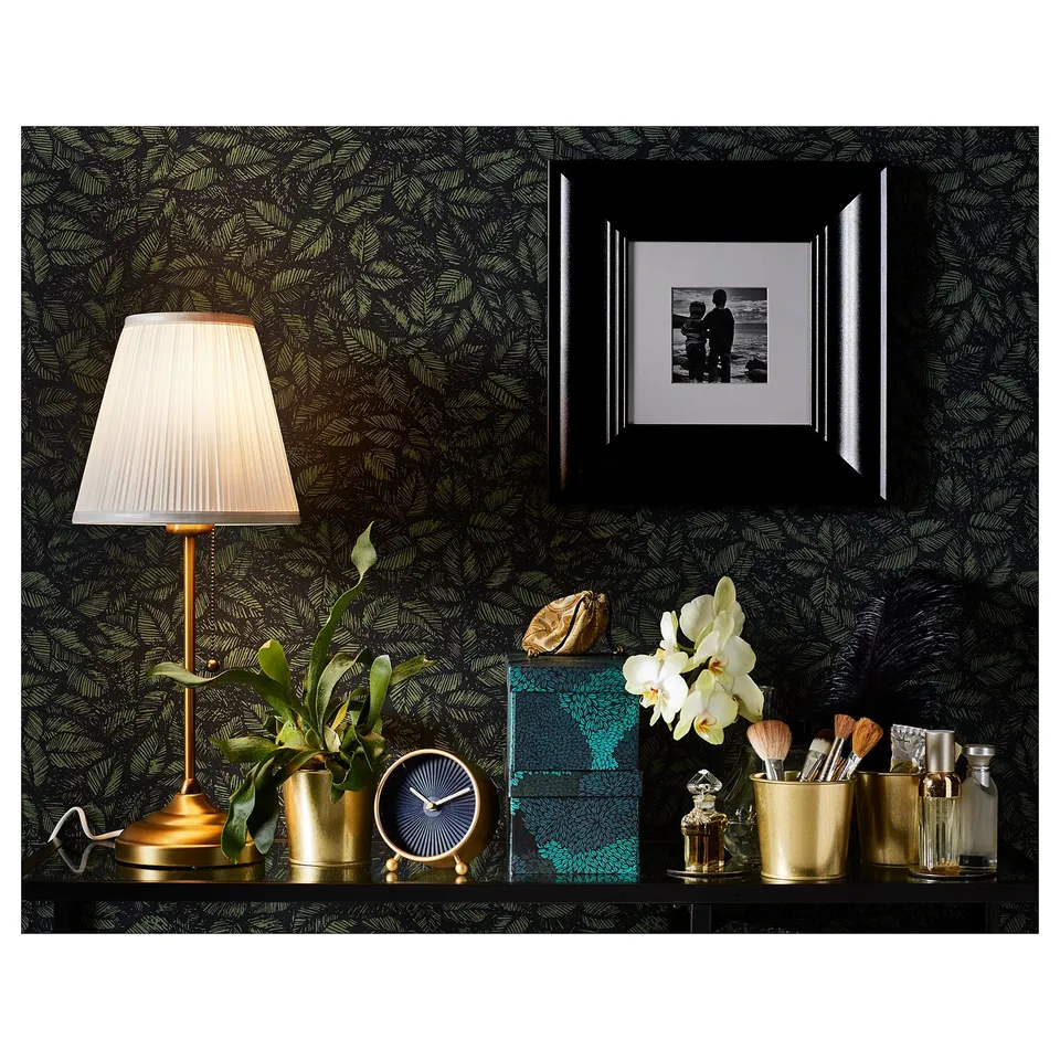 Elegant desk setup: black lamp, gold clock, green box, white flowers, makeup brushes, photo frame against dark leafy wallpaper.