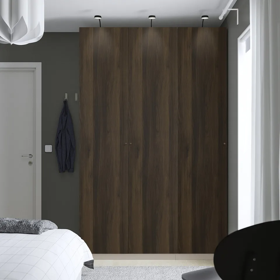 Modern bedroom large wooden PAX wardrobe, dark wood finish, spanning wall height. A white bed and grey wall with hooks are nearby.