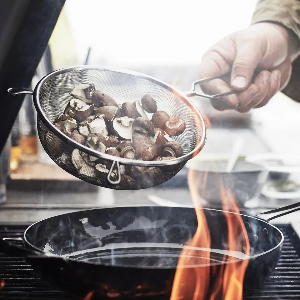 Chef holding sieve with mushrooms over sizzling skillet.