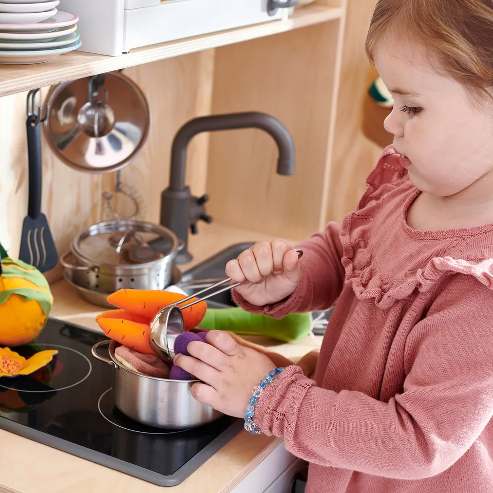 Child playing with a silver DUKTIG toy stove, pretending to cook vegetables.