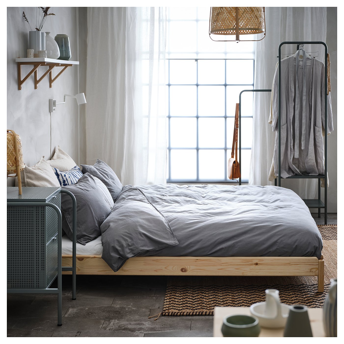 Bedroom with a grey comforter on a wooden bedframe near a window with sheer curtains. Shelf with vases and clothes rack with grey clothing.