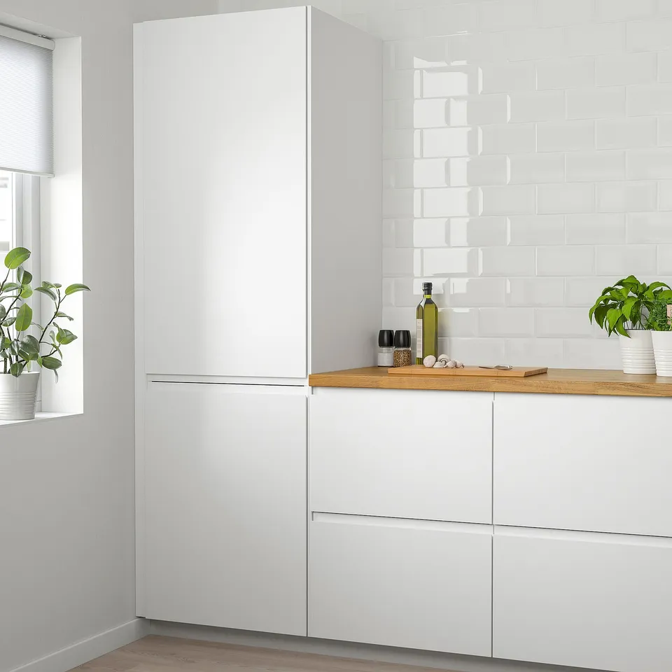 Modern white kitchen with tall VOXTORP fridge, exposed wood counter, plants, and white brick backsplash.