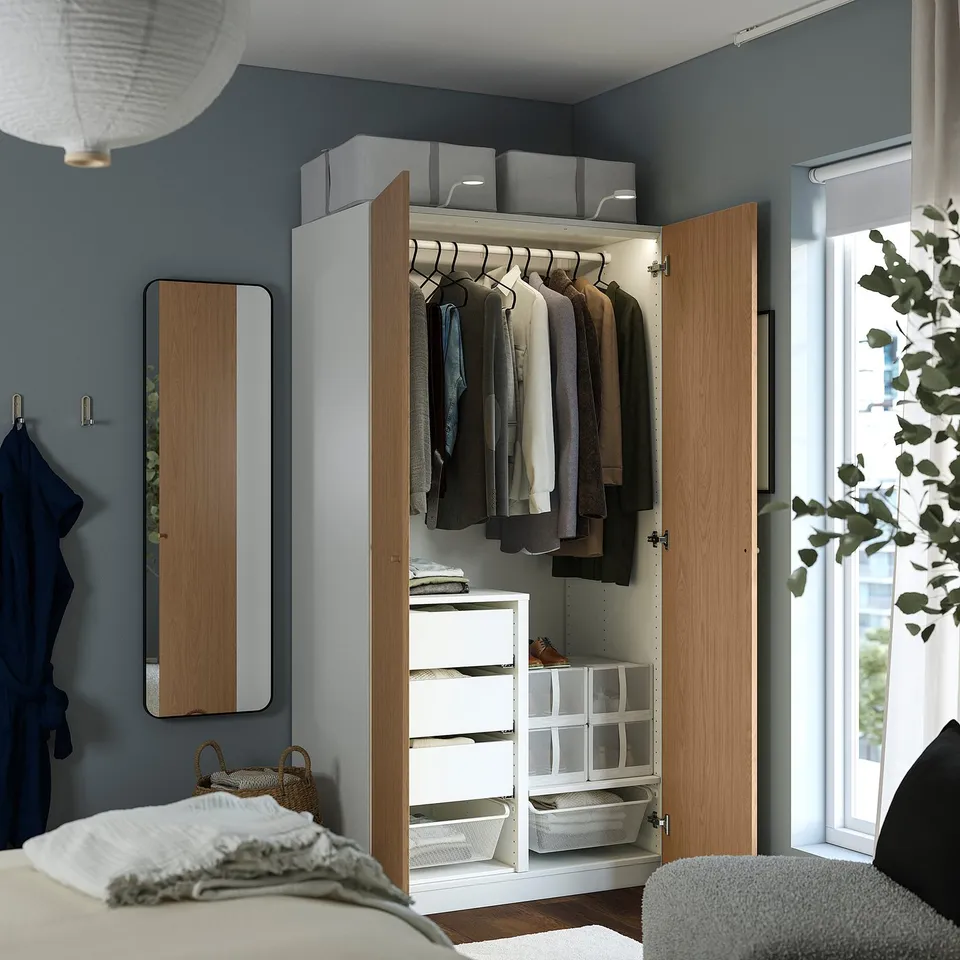 Bedroom with open wooden PAX wardrobe showing clothes, drawers, and storage boxes.