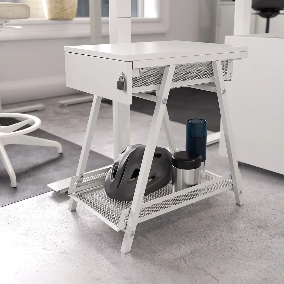 White TROTTEN storage unit with lockable drawer, mesh shelf, and a-shaped frame, holding a black helmet and blue cup.