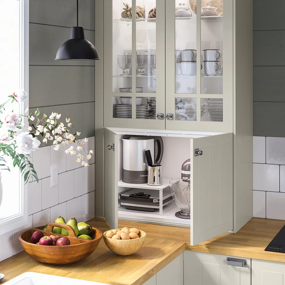 Kitchen with light grey glass-front cabinets, white counter, wooden bowl with fruits, and pendant lamp.