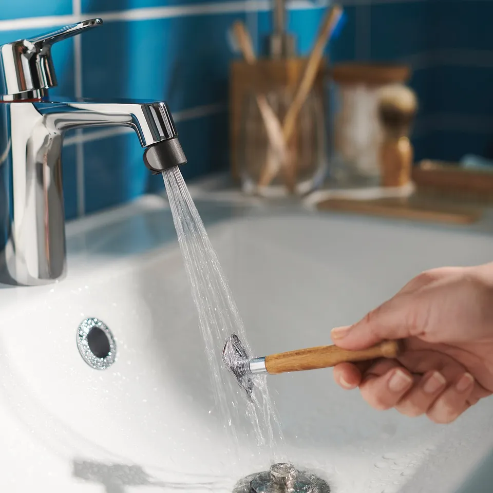 A person washing a toothbrush under a modern silver tap with a chrome ÅBÄCKEN nozzle in a blue-tiled bathroom.