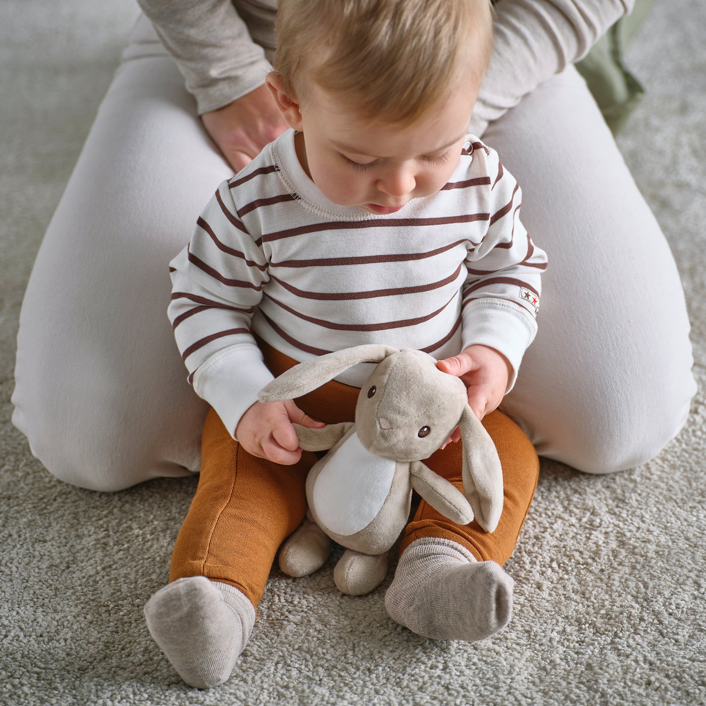 A baby sits hugging a plush bunny toy between legs. The toy has long ears, is soft, and is designed for cuddling and play.