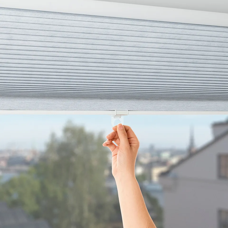 A hand adjusts a cordless, honeycomb-style block-out blind, illuminating a bright, cityscape view.