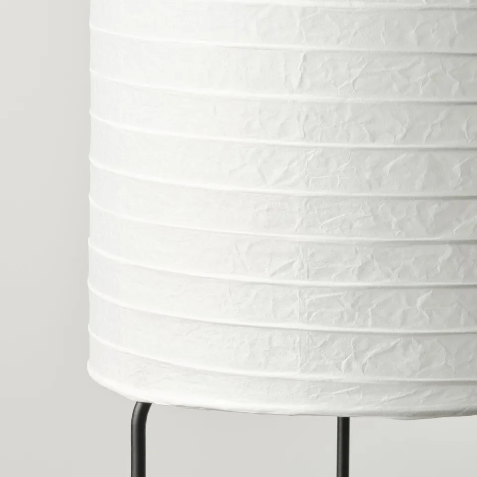 Cylindrical lamp with textured white rice paper shade supported by metal legs. Elegant and minimalist design, emitting soft diffused light.