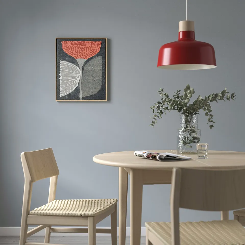 Abstract art BLODFLÄDER above dining table with wicker chairs, red lamp, vase with foliage, glass, and newspaper.