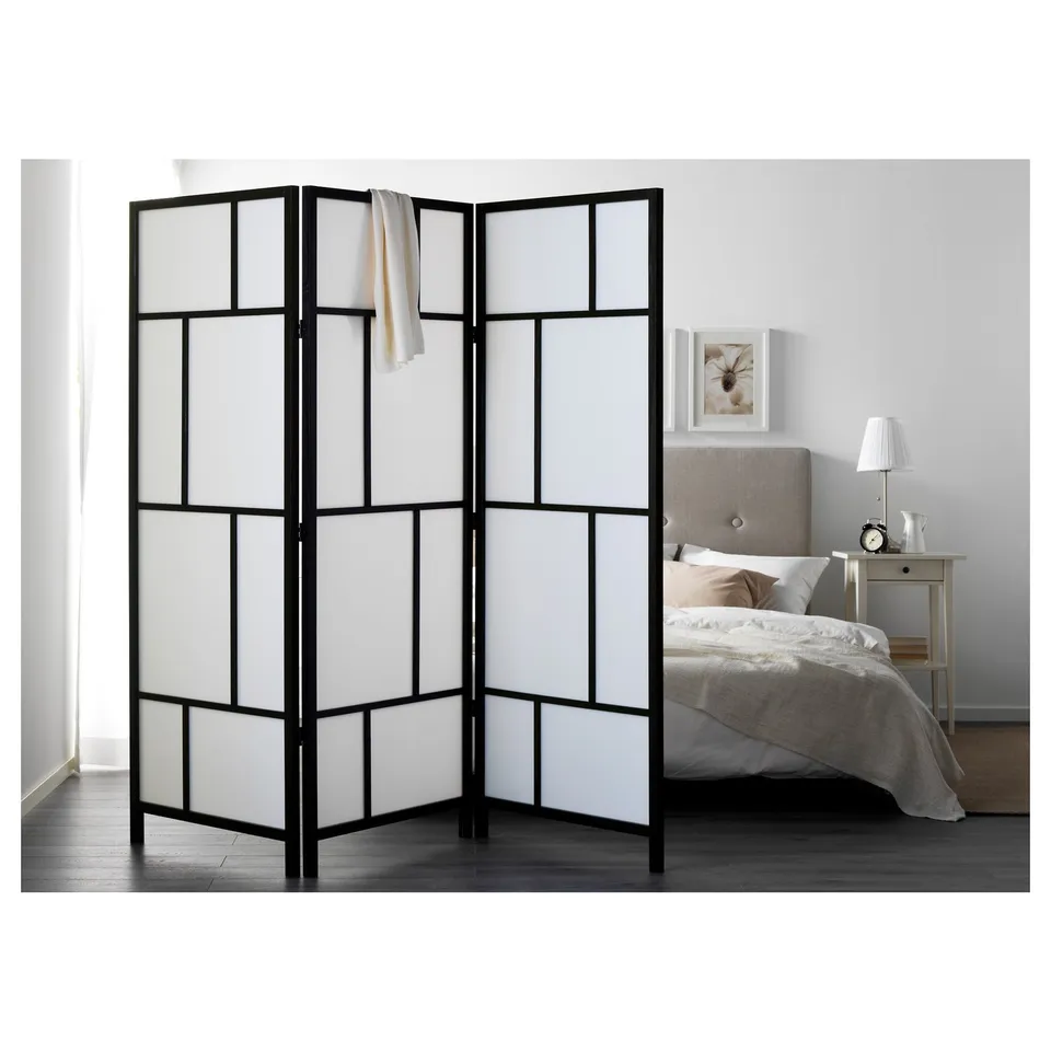 Black & white pine RISÖR room divider with grid design, larger than bed & bedside table, draped with cloth.