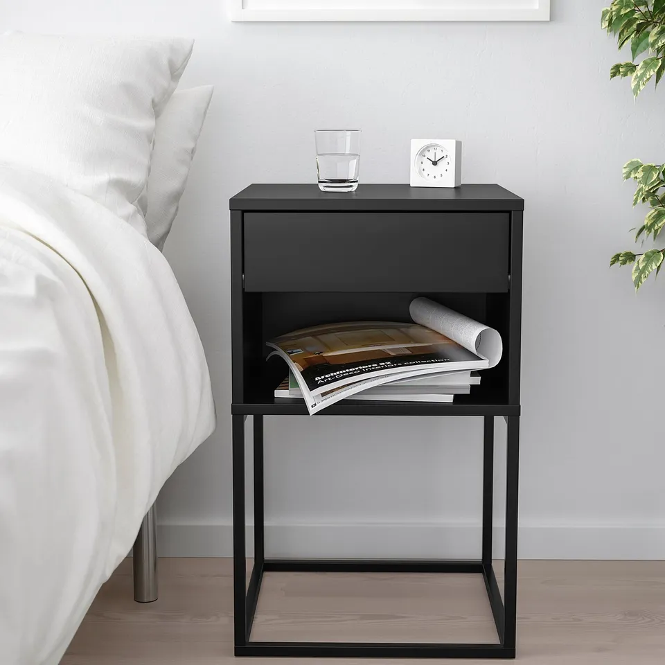Black VIKHAMMER bedside table beside bed with drawer and shelf.