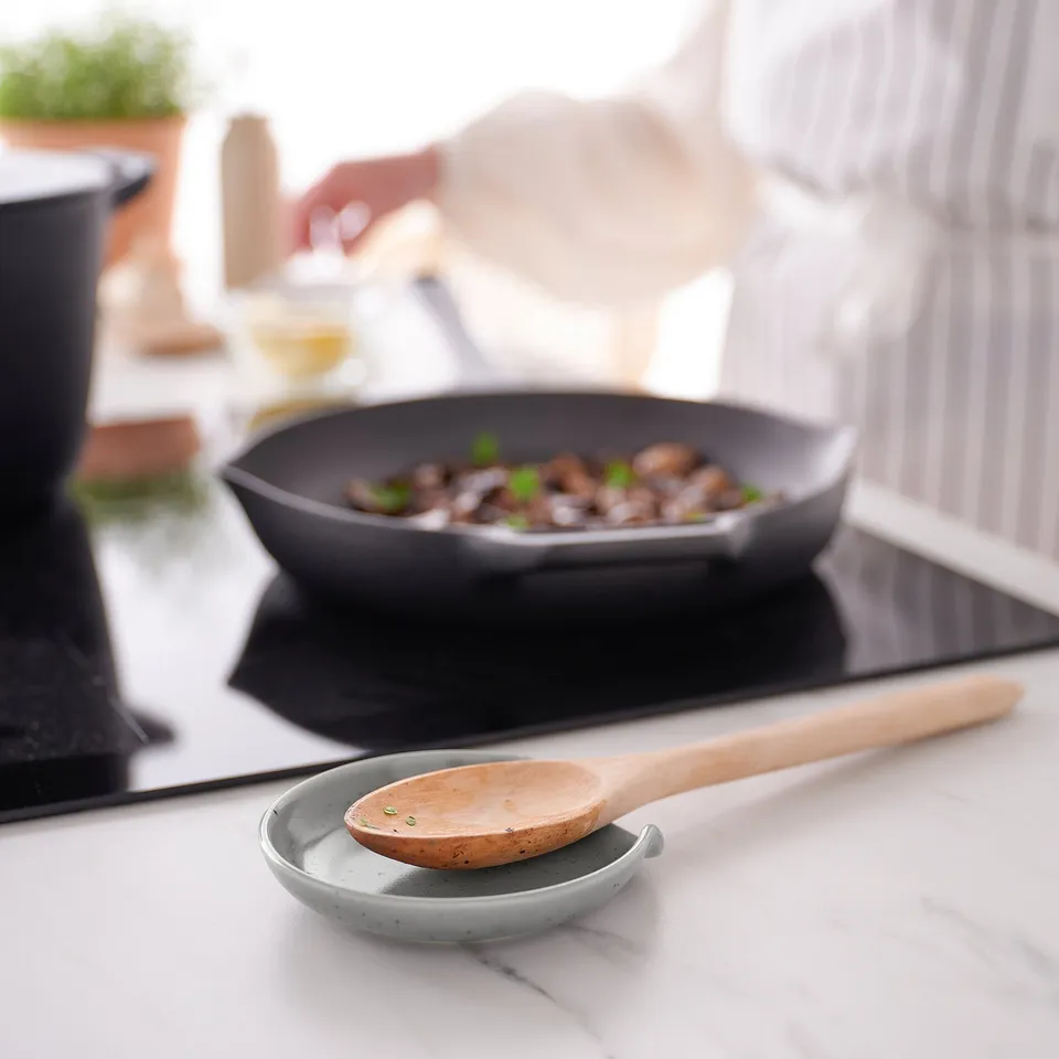 A spoon rest on a countertop, holding a wooden kitchen spoon. Behind it, a pan of food cooks on the stove.