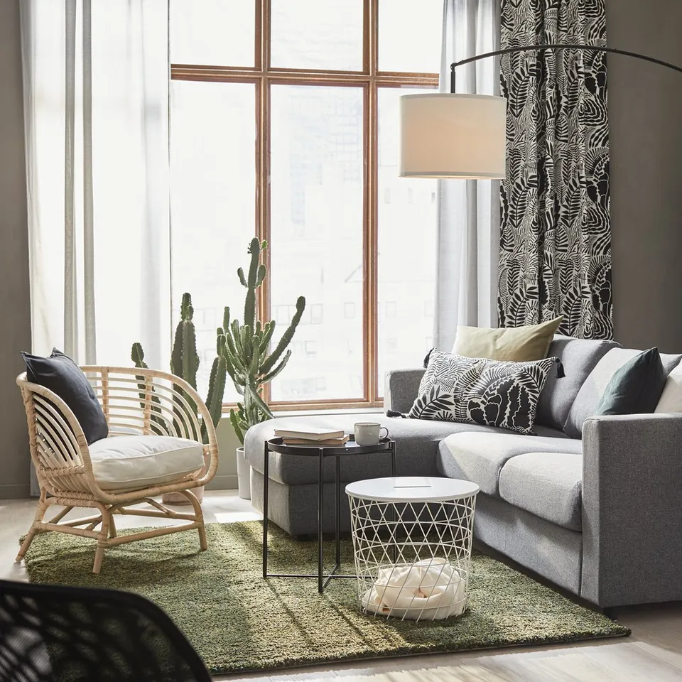 Modern living room with grey VIMLE sofa, black cabinet, and large window. A green rug, wicker chair, and floor lamp.