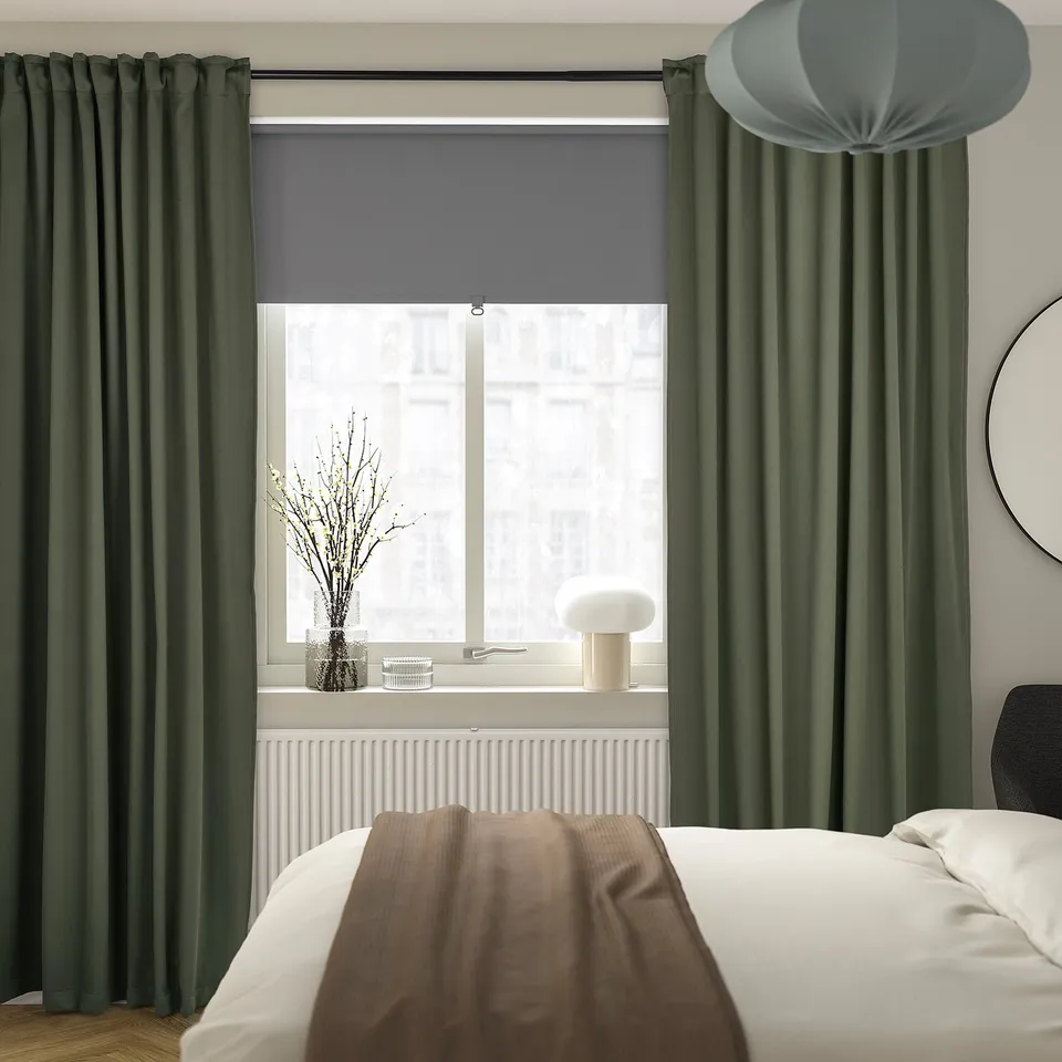 Modern bedroom with MAJGULL dark green block-out curtains. White bed with brown throw, grey lamp, and green curtains.