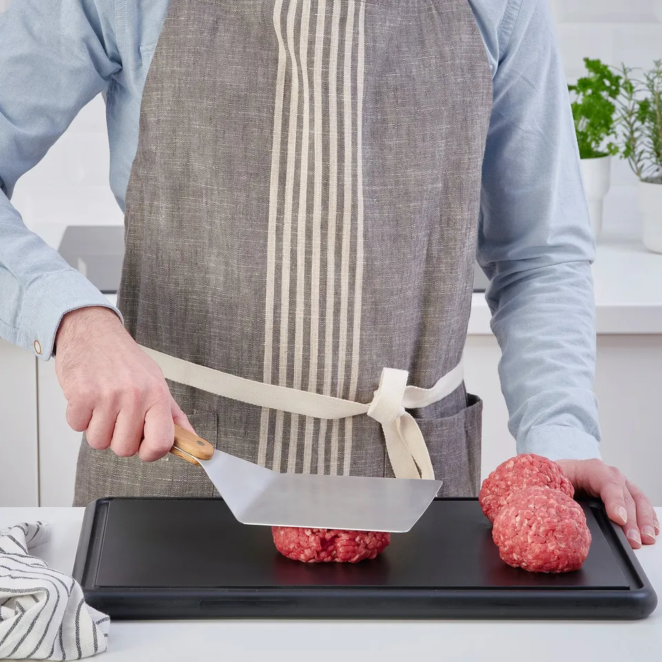 Person using a wide spatula to flip burgers on a tray. They wear an apron and striped shirt. The spatula is wooden-handled, ideal for grilling.