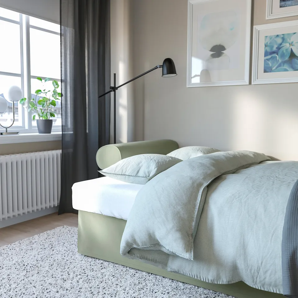 Modern bedroom with large green sofa-bed under a wall-mounted black lamp, clean white walls, large windows with dark curtains, and a white radiator.
