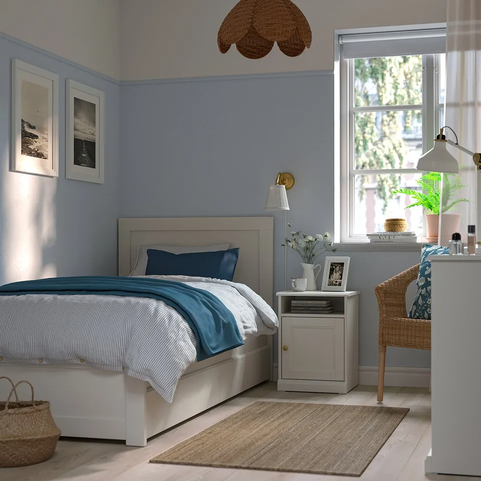 Cosy bedroom with grey FÅGELFJÄLLET bedside table, flowers, lamp, wicker chair, large window, blue linens, artwork, and wicker pendant light.