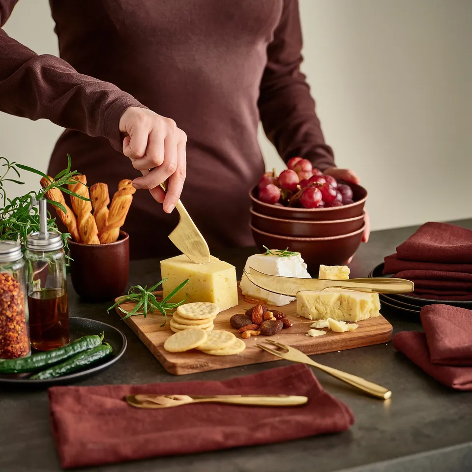 Person cutting cheese on a wooden board with crackers, almonds, and grapes on a table with elegant brass-colored serving utensils.