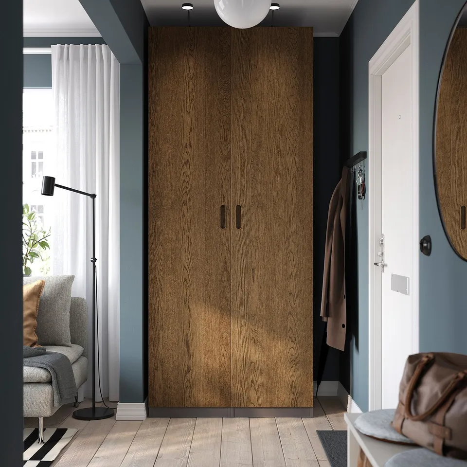 A modern bedroom with a tall brown TONSTAD wardrobe, white door, grey couch, and coat rack, well-lit by a floor lamp.