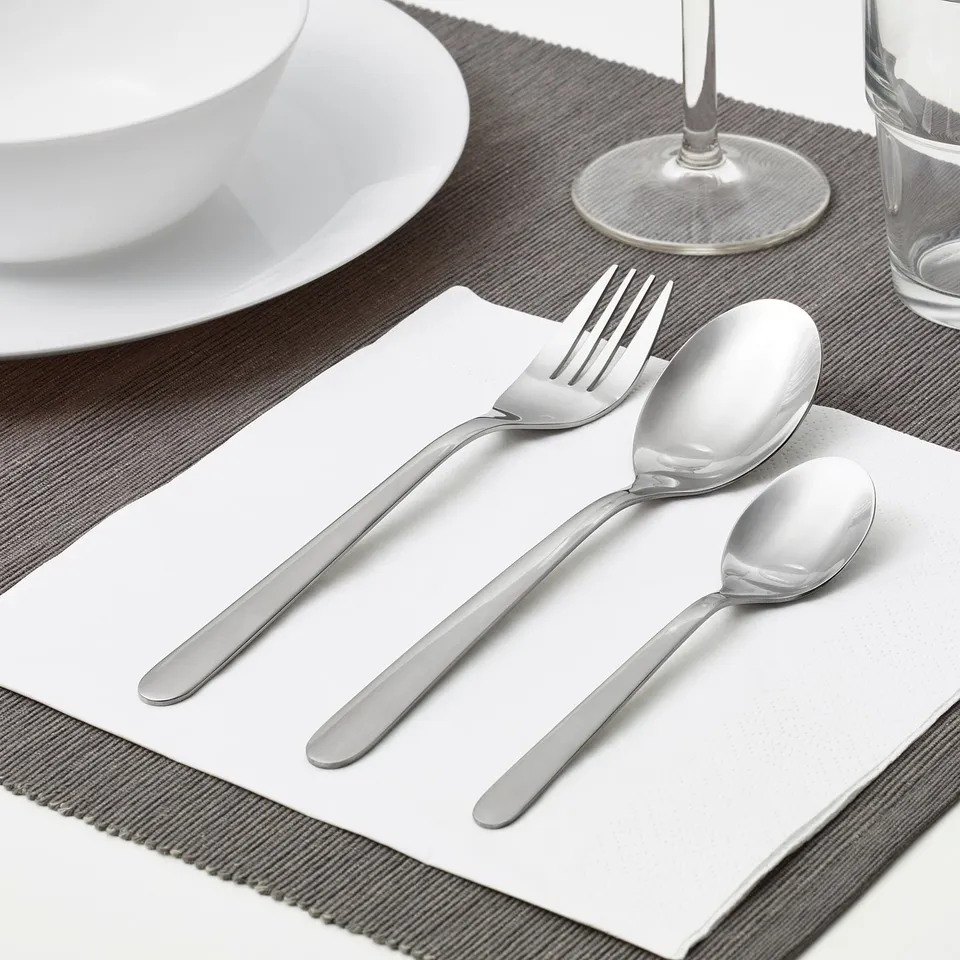 A neatly arranged table setting featuring a fork, spoon, and teaspoon on a napkin, alongside a plate and glass, showcasing everyday cutlery.