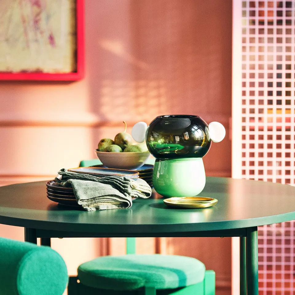 Small green IKEA table with metal legs and round shape, holding stacked green vases, a bowl of fruits, and plates, near green chairs.