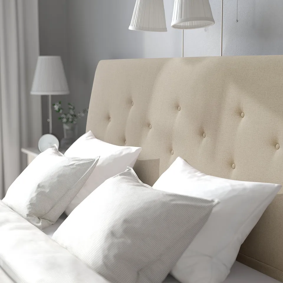 A beige upholstered bed with a sloping headboard and multiple white pillows. Overhead lamps with white shades beside the bed.