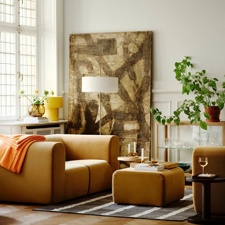 Living room with golden sofa, tall lamp, plants, art.