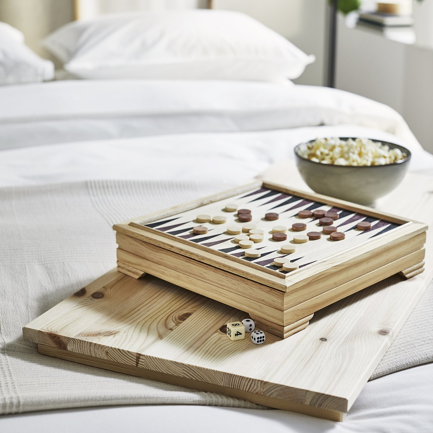 Wooden backgammon set on bed; includes dice and bowl of snacks.