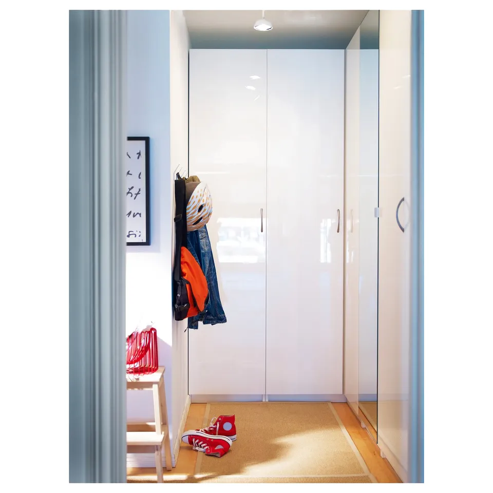 White glossy closet doors with modern design, hanging coats, red trainers, blue bag, light wooden floor, art frame, light blue curtain.