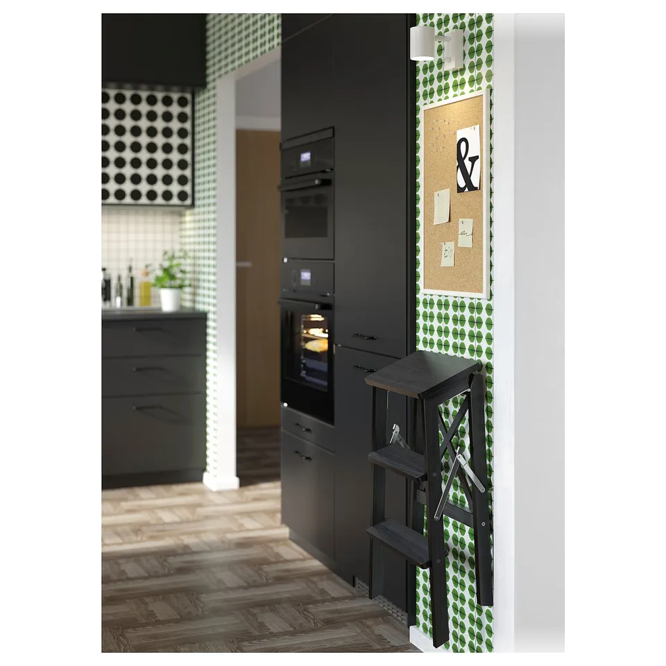 Modern kitchen featuring black cabinets, built-in oven, step ladder, green patterned wallpaper, and a corkboard with notes.