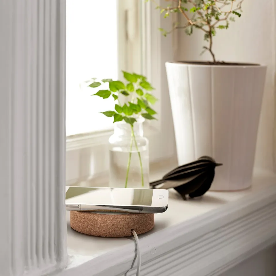 White phone on circular cork wireless charger by window with plants.