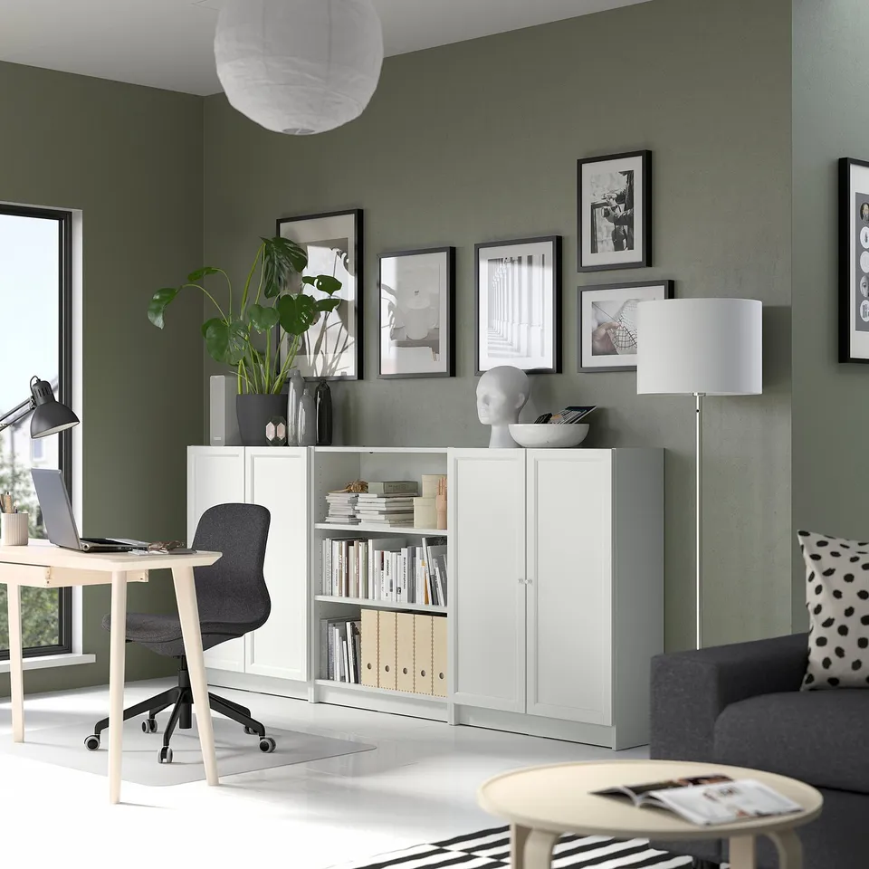 Modern home office with white BILLY bookcase, grey sofa, and desk. Green walls, art, and plants decorate the space.