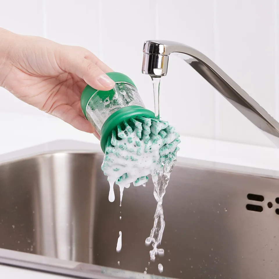 Person holds dish brush under sinks tap, dispensing soap for washing.