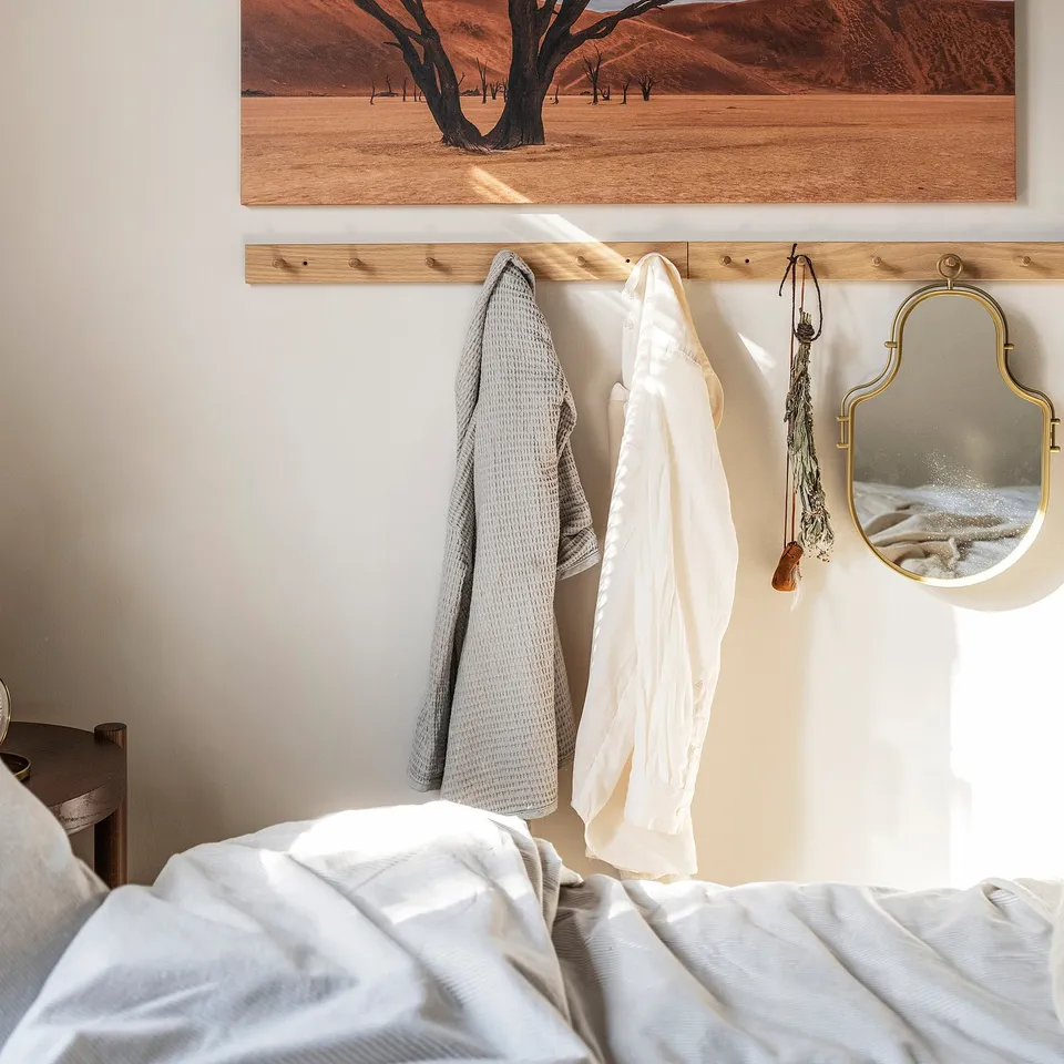 Bedroom wall with HÖVOLM oak coat rack holding white and grey robes, small mirror, and desert landscape.