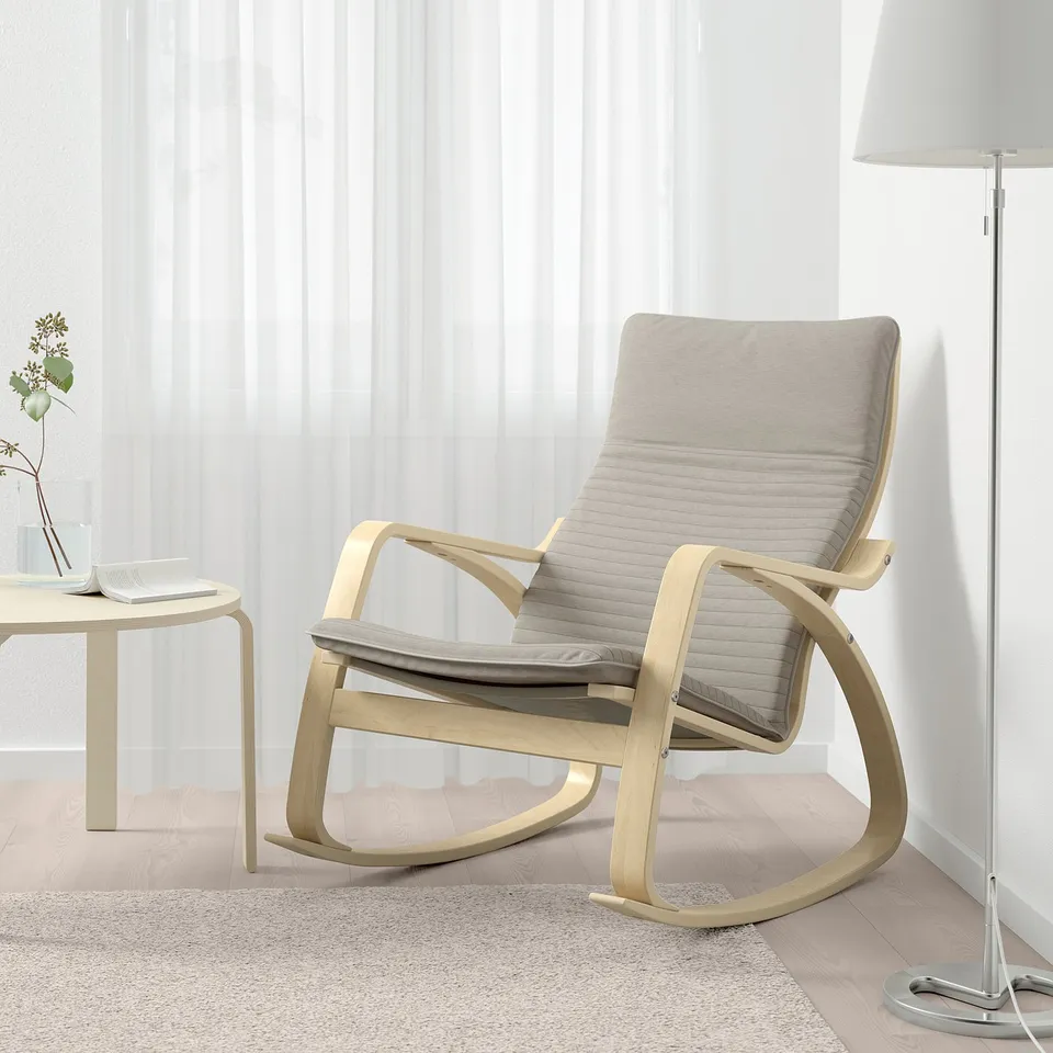 A light beige rocking chair with a cushioned seat and back on a light wooden floor, beside a small table with a plant and a tall lamp.