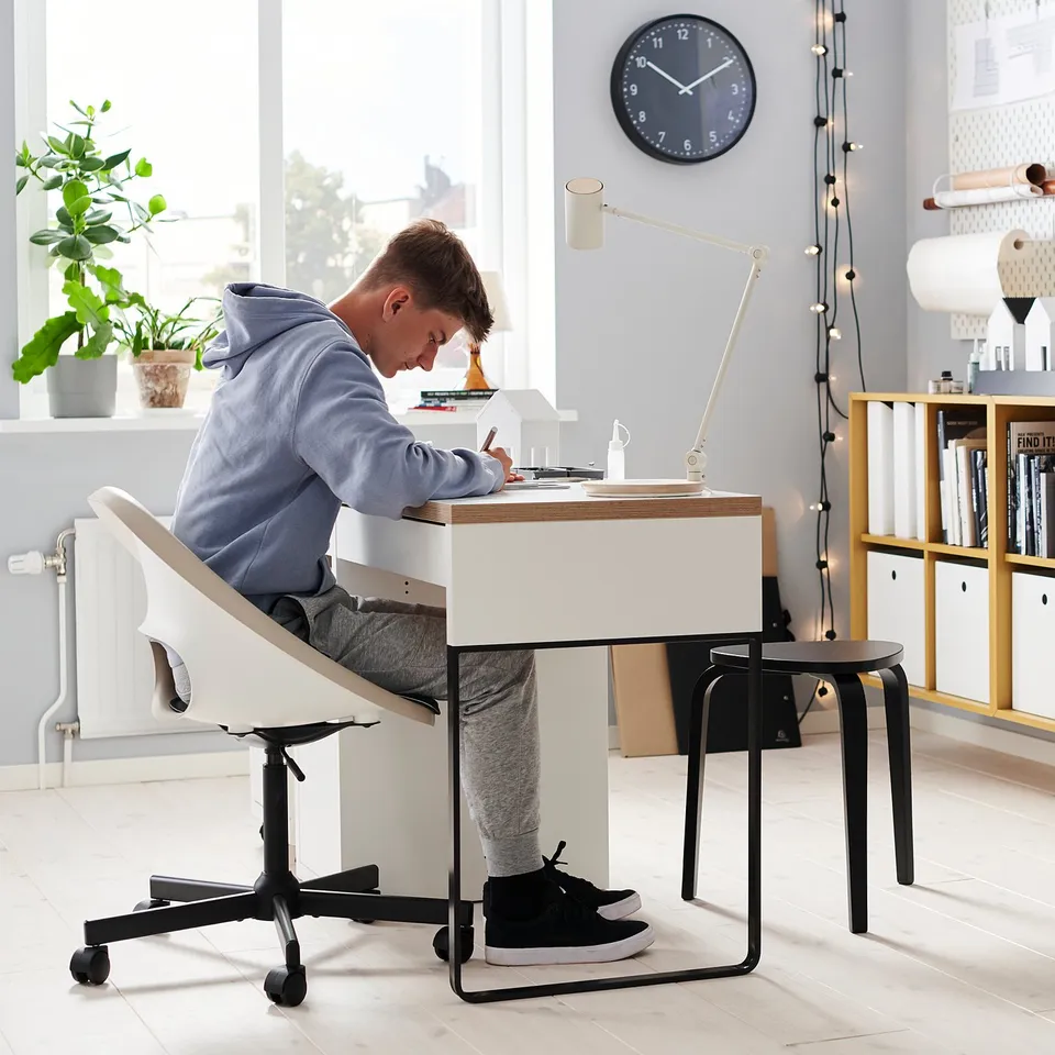 Person sits at ELDBERGET desk chair, light blue hoodie, sketching on white desk. Modern, clean workspace with light wood and black accents.
