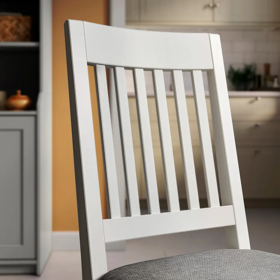 A close-up of a white wooden chairs backrest featuring vertical slats, set in a modern kitchen. The chair has a sleek, minimalist design.