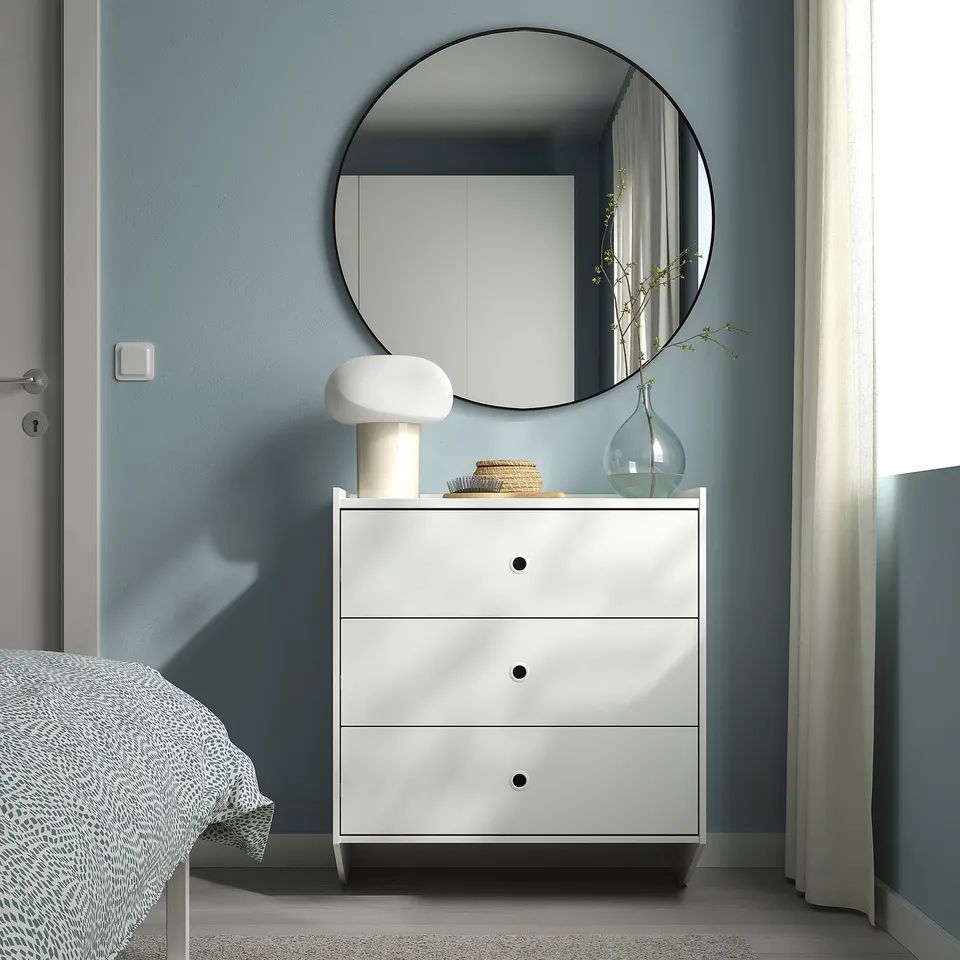 Modern bedroom: RACKNÄS white chest in corner, blue wall backdrop, mirror, lamp, vase with branches.