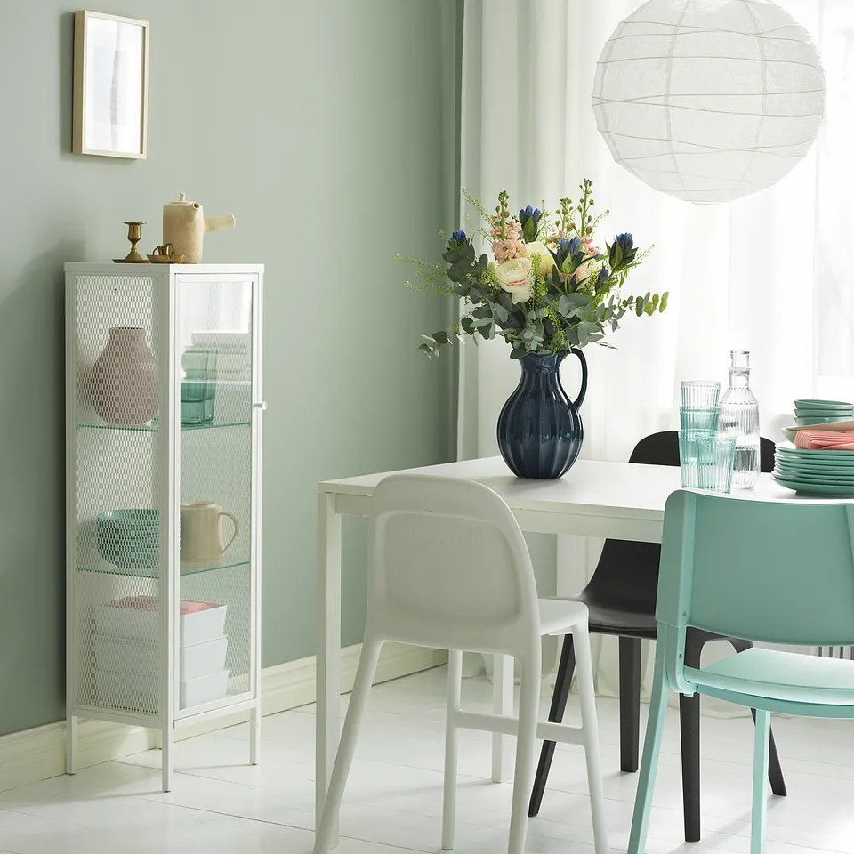 Modern dining room with light green walls, white table, BAGGEBO cabinet, blue vase, light fixtures, and chairs in green, white, black.
