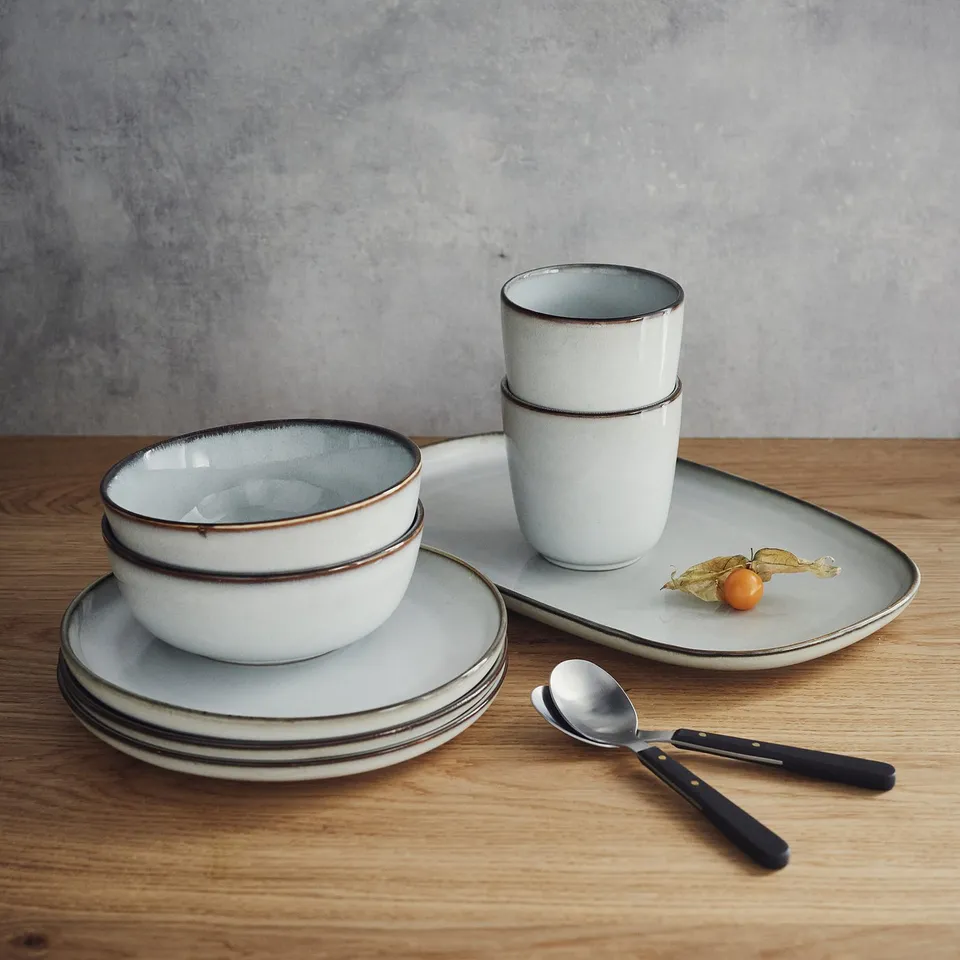 GLADELIG tableware: white stoneware dishes, bowls, cups with rustic, sandy glaze. Stacked, with a spoon and fruit on table.