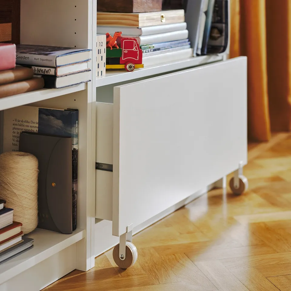 White BILLY bookcase with drawer and wheels, holding books and toys, on wooden floor.