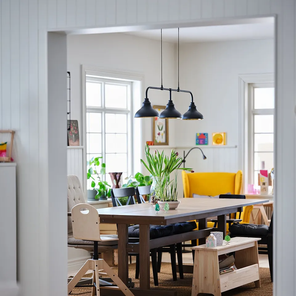 Bright, stylish dining area with PERJOHAN wooden bench, chandelier, yellow chairs, table with vase.