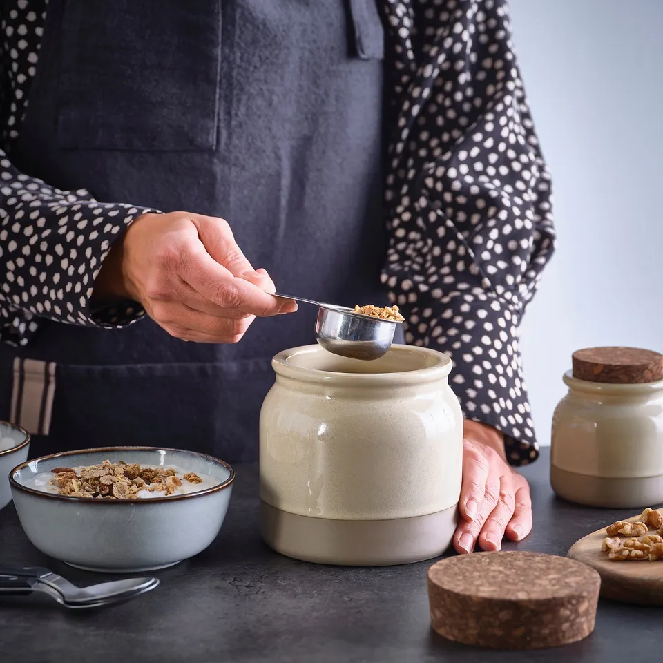 A person scoops granola from a stoneware jar with a cork lid, displaying its practical storage use.