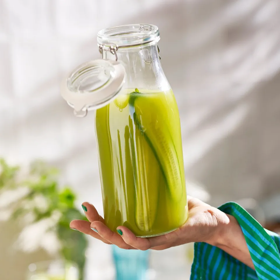 Hand holding glass jar with green liquid and cucumber, sealed with white lid.