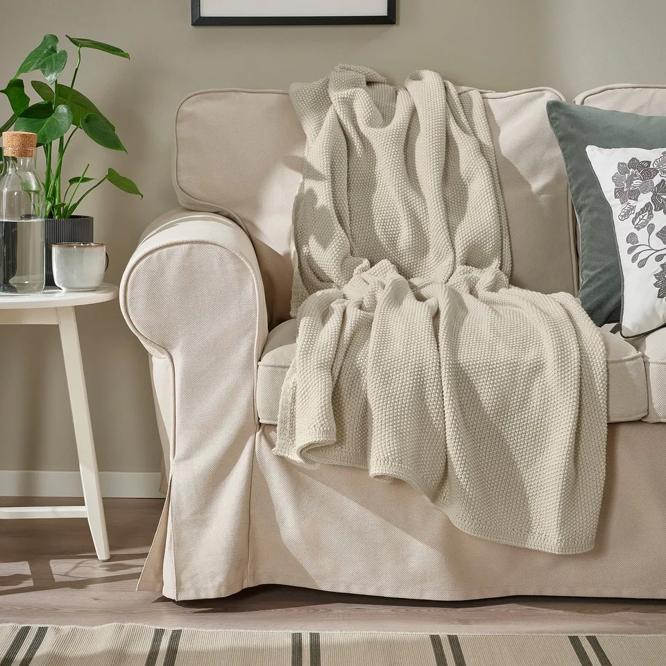 Beige couch with HUMLEMOTT throw blanket, soft, recycled polyester, off-white, cosy. Decorative side table, plant, mug, pillow.