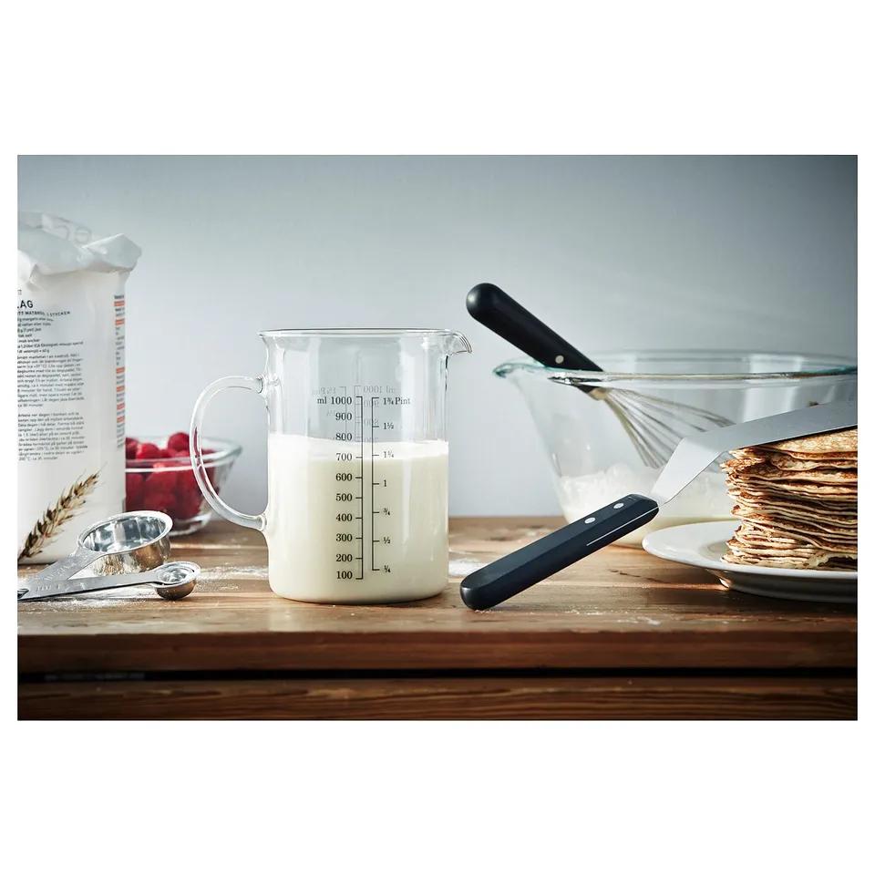 Clear glass measuring jug with black handle, filled with cream. Marked with measurements and placed on wooden table with baking tools.