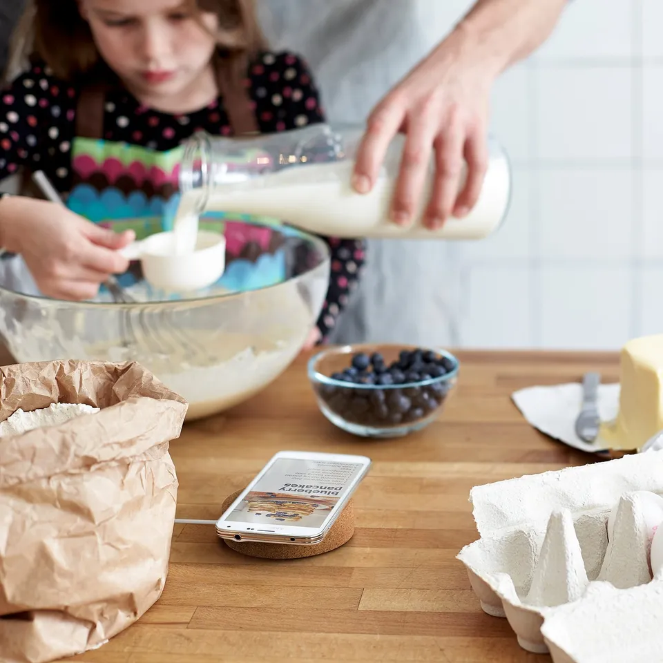 Child baking with adult, using smartphone on wireless charger, ingredients nearby.